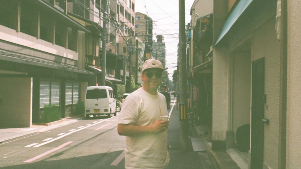 faded photo of a man standing near a city street smiling at the camera with sunglasses on